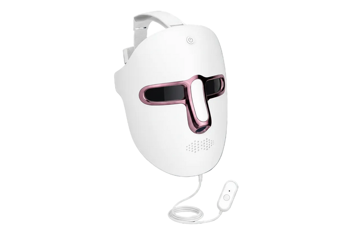 A white LED facial therapy mask with a pink metallic accent around the eye cutouts, shown with its attached power cable and remote control against a dark gradient background.